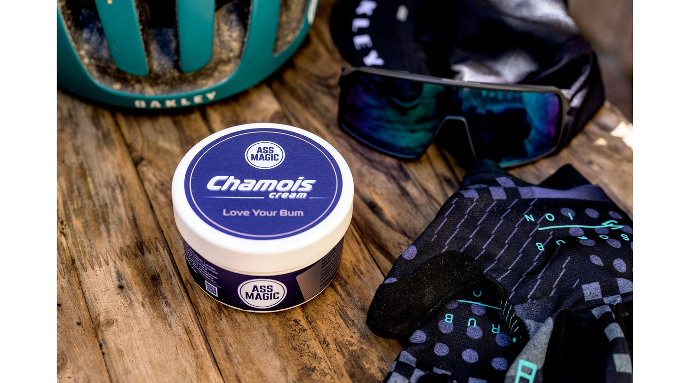 ASS MAGIC Chamois Cream - 200ml Tub laying next to various cycling accessories (cycling sunglasses and gloves)