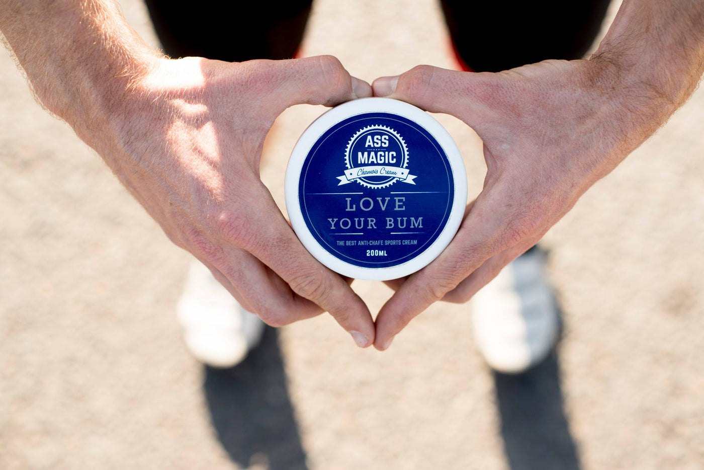 ASS MAGIC Chamois Cream Tub being held by a male's hands in the shape of a heart with text reading "Love Your Bum" on the label